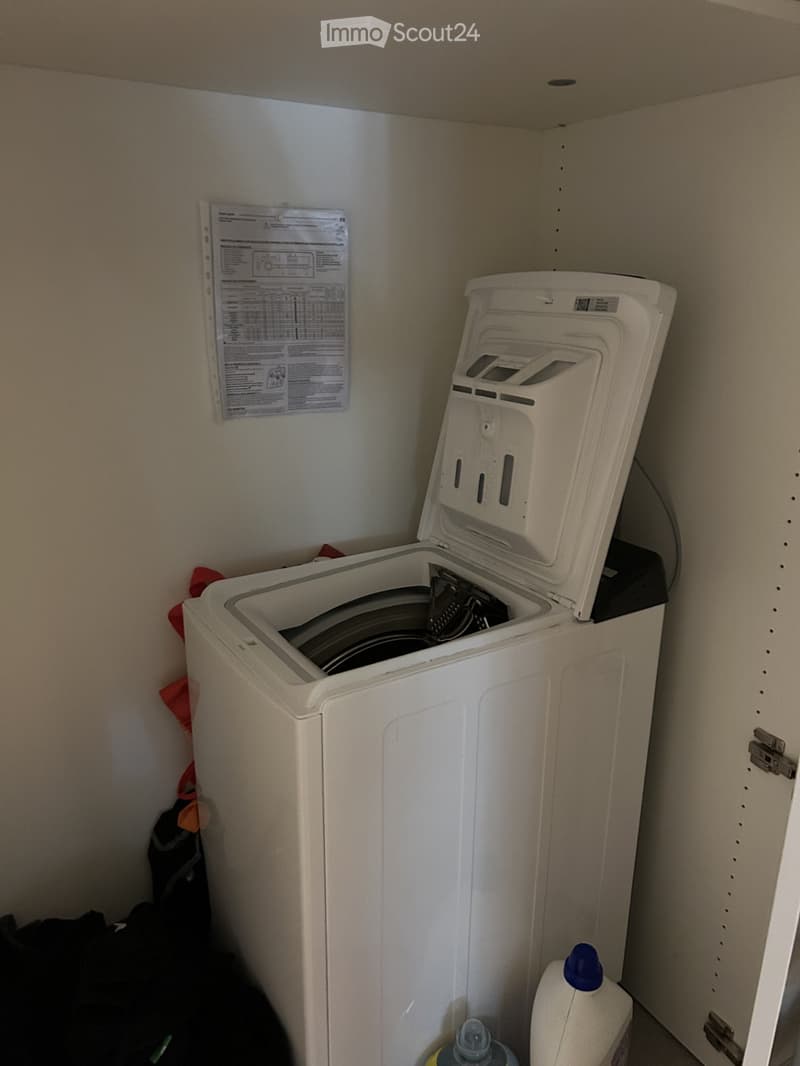A compact laundry area with a white top-loading washing machine, detergent bottle, and instruction manual on the wall.