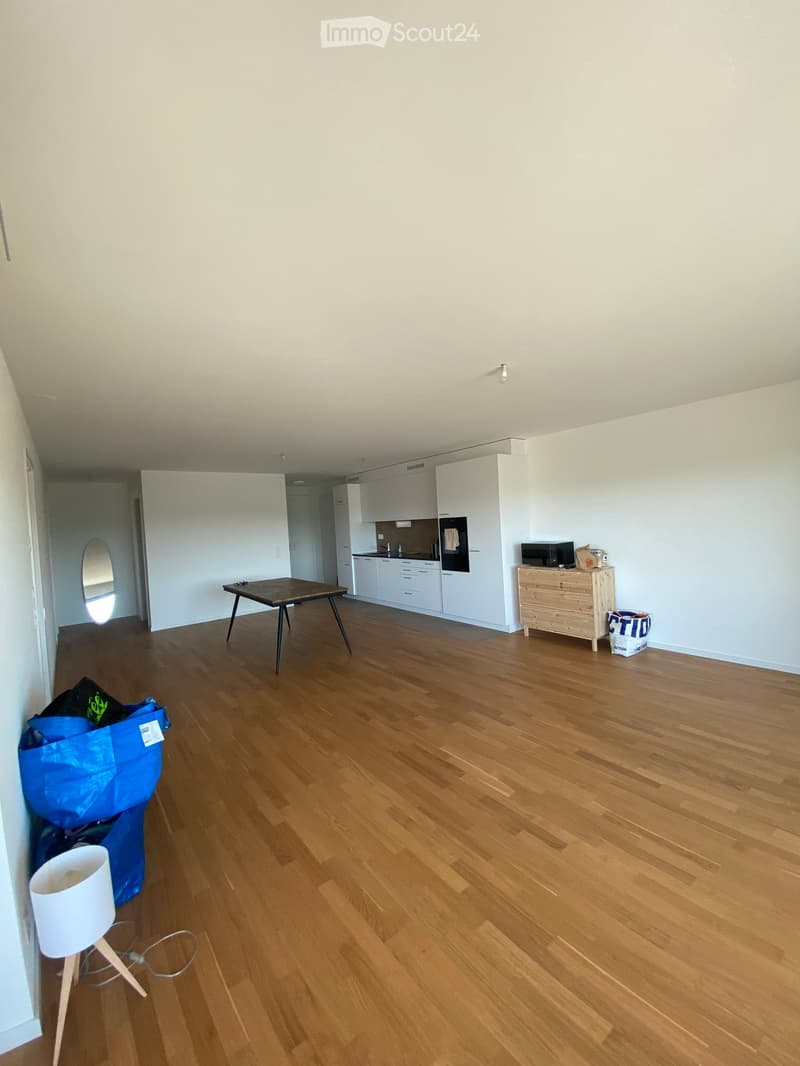 A partially furnished room with wooden flooring, a small table, a wooden cabinet, a microwave, and a lamp. The room is connected to a kitchen area with white cabinets and a countertop.