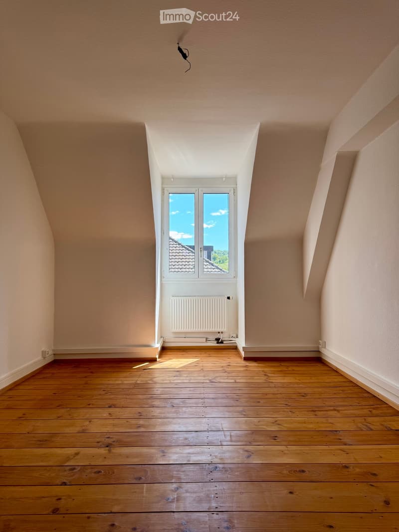 Empty attic room with wooden flooring, slanted ceilings, and a large window offering a view of the roof and sky.