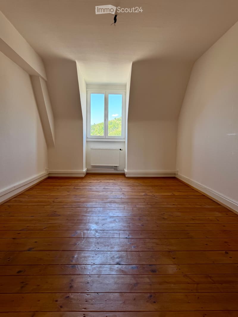 Empty attic room with wooden flooring, white walls, and a single window with a radiator below it.