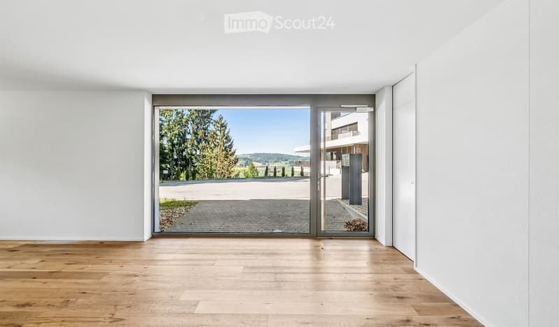 Empty room with wooden flooring and white walls, featuring a large glass door leading to an outdoor area with trees and a paved driveway.