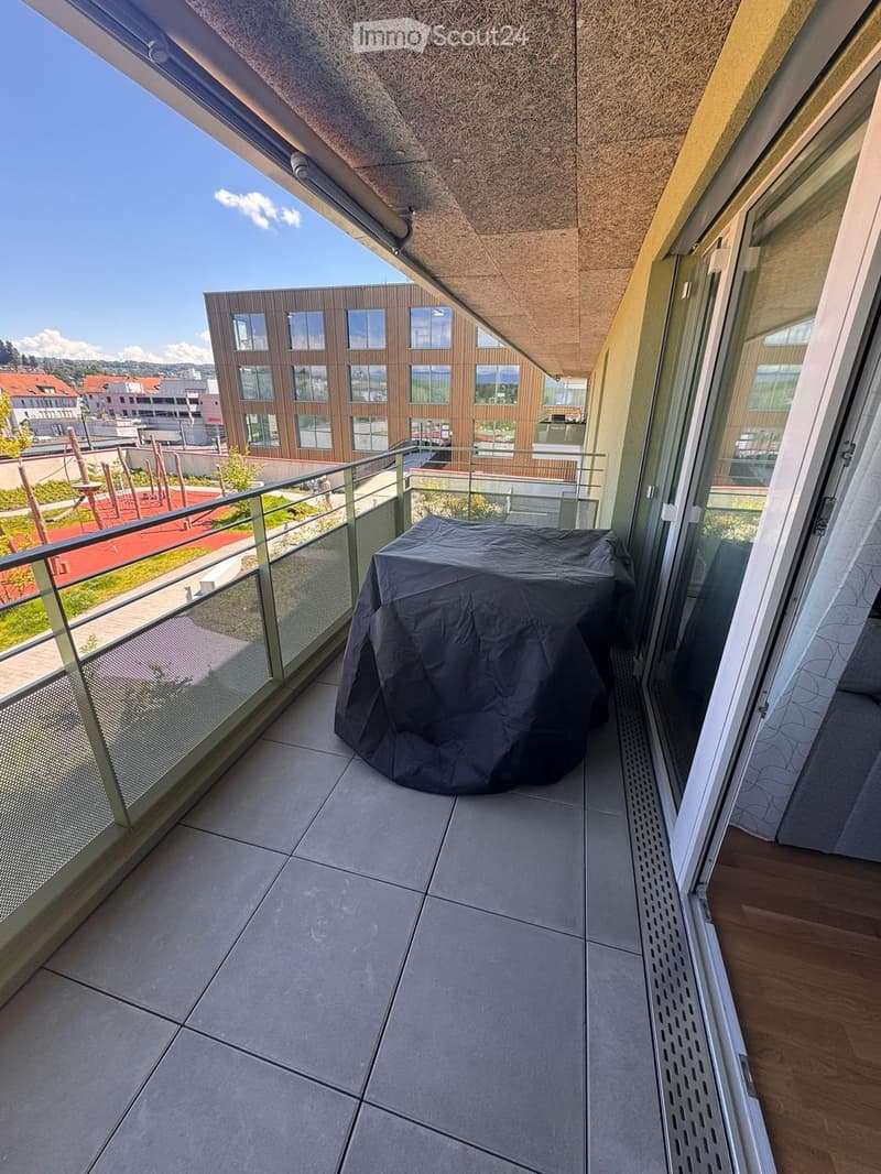 Balcony with tiled floor, glass railing, and covered outdoor furniture, overlooking a modern building and playground area.