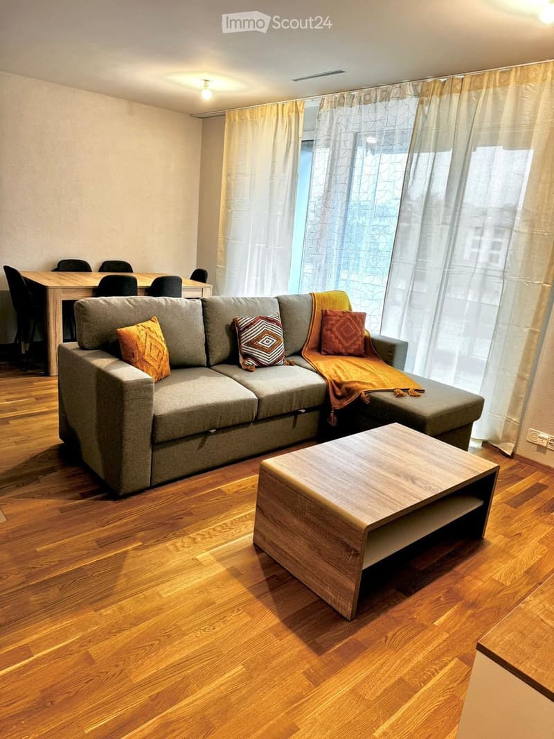 Modern living room with a grey sectional sofa, wooden coffee table, dining table with chairs, and large windows with curtains.