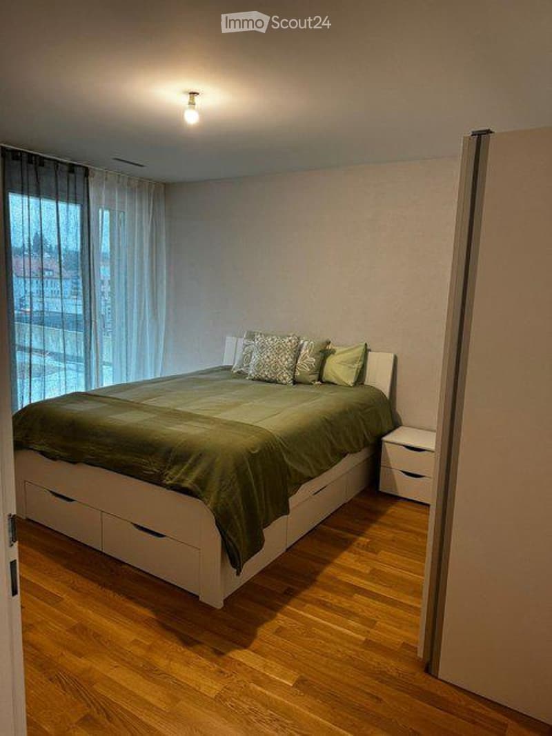 Modern bedroom with a white bed frame, green bedding, and a nightstand. Large windows with sheer curtains allow natural light to fill the space.