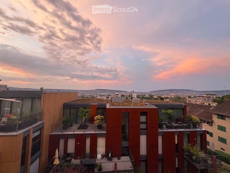 Modern multi-story residential building with balconies, greenery, and cityscape view in the background.