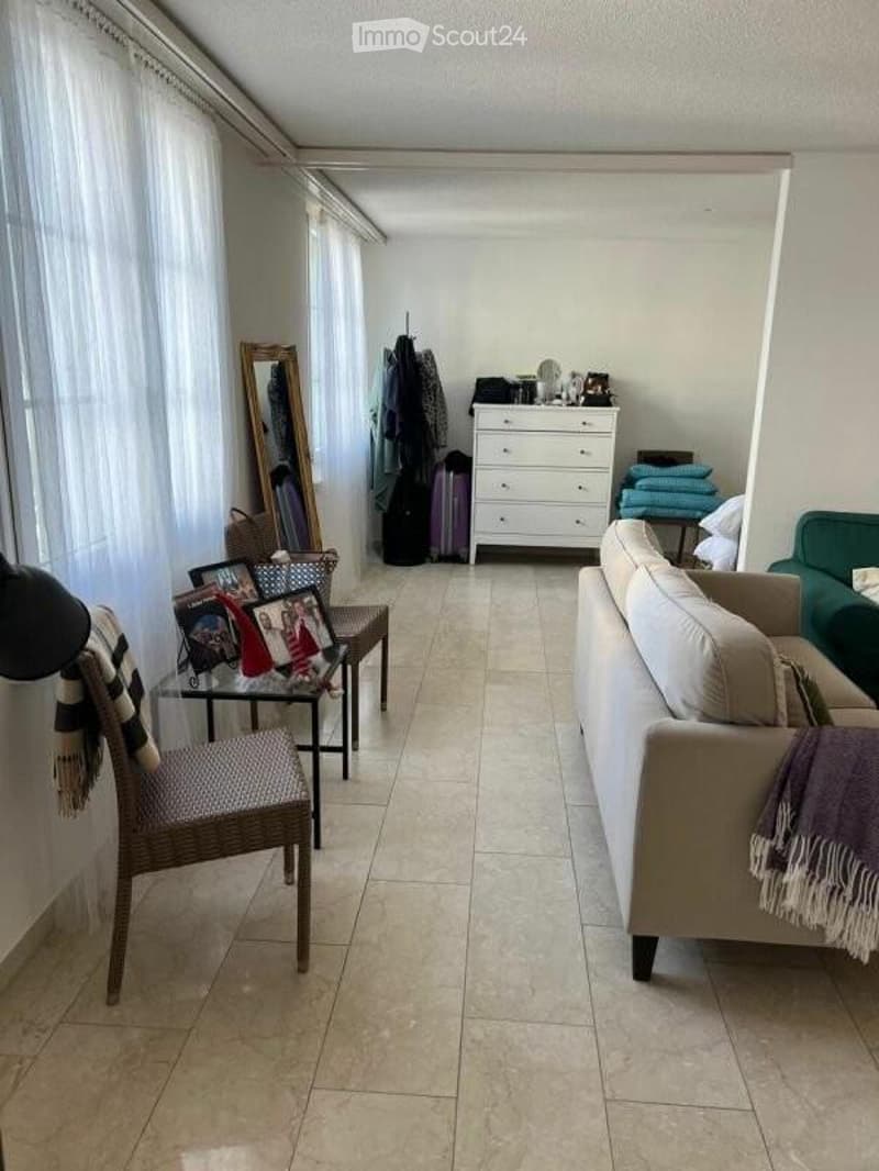 A well-lit living room with a beige sofa, a wicker chair, a glass coffee table, a dresser, and a mirror. The room features large windows with white curtains and a tiled floor.