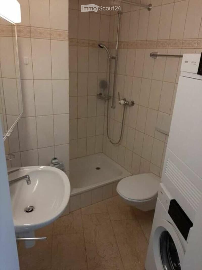 A compact bathroom with a white sink, toilet, shower, and a washing machine. The walls are tiled in light beige, and the floor is covered with beige tiles.