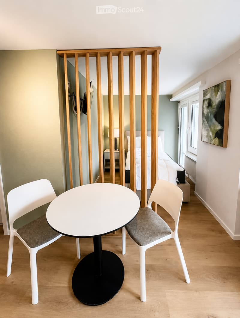 Modern, minimalist room with a round white table, two white chairs, a bed with white bedding, a nightstand with a lamp, and a painting on the wall.