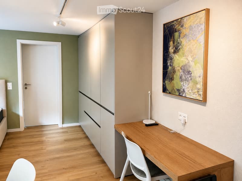 Modern, minimalist room with light wood flooring, white walls, a tall cabinet, a desk with a chair, and a painting on the wall.