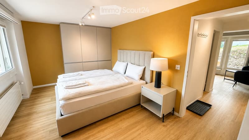 Cozy bedroom with a double bed, nightstand, lamp, wardrobe, and a chair near the window.