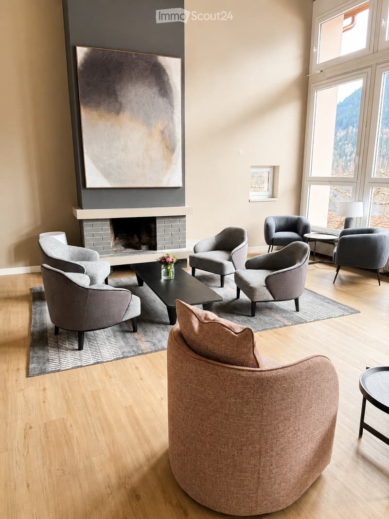 Modern living room with large windows offering a scenic view, a fireplace, grey armchairs, a sofa, coffee table, and a side table with a lamp.
