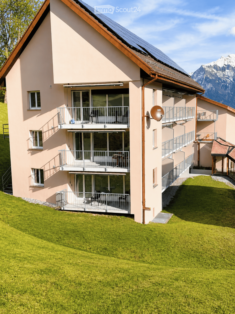 A two-story residential building with light pink walls, multiple balconies, and a green lawn in front. The building features large windows and a solar panel on the roof, with a scenic mountain view in the background.