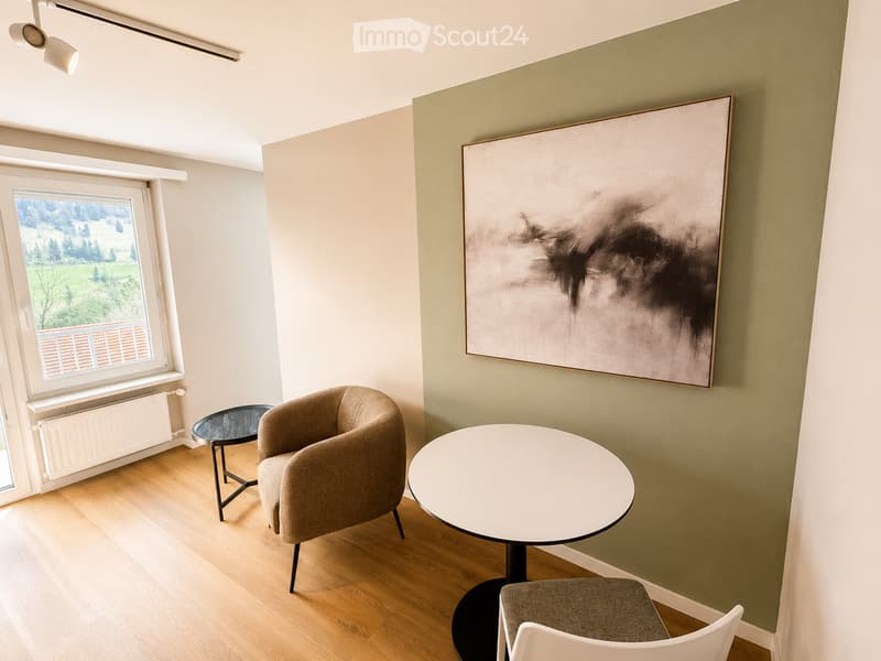 A modern, minimalist room with light wooden flooring, a large abstract painting on the wall, a round white table, a beige armchair, a small black side table, and a white stool.