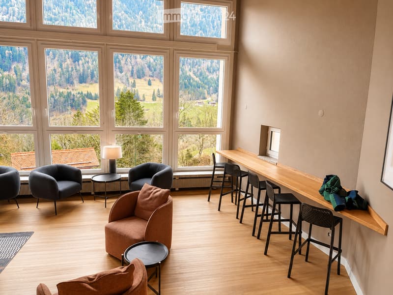 Bright, modern living space with large windows offering a scenic mountain view. Features include a wooden floor, a long wooden counter, several chairs, a sofa, and a lamp.