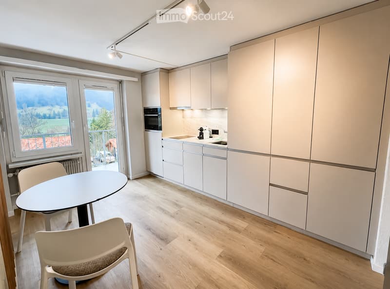 Modern kitchen with light-colored cabinets, a round dining table with two chairs, and a view of a balcony through large windows.
