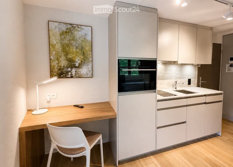 Modern kitchen with white cabinets, a wooden desk, a white chair, a sink, and an oven. A lamp and a picture frame are also visible.