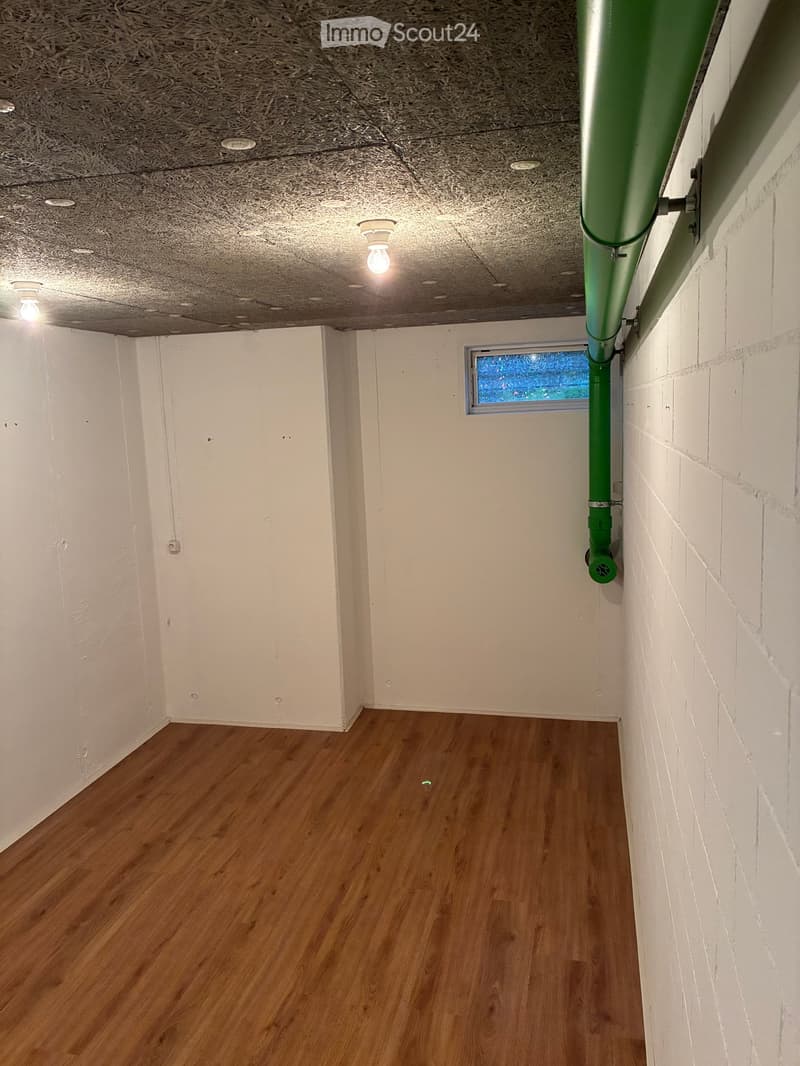 Empty hallway with wooden flooring, white walls, and a green pipe on the right side. A small window is visible at the end of the hallway.