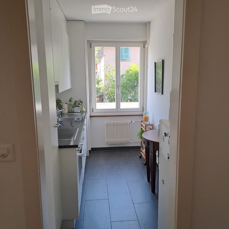 Compact kitchen with tiled floor, white cabinets, sink, fridge, radiator, small table, and potted plants.