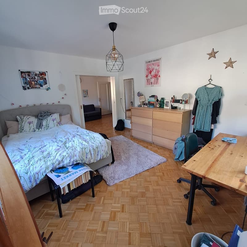 Cozy bedroom with a wooden floor, a large bed with a floral duvet, a wooden desk with a chair, a small sofa, a dresser, and a mirror. The room features a hanging pendant light and several decorative items on the walls.