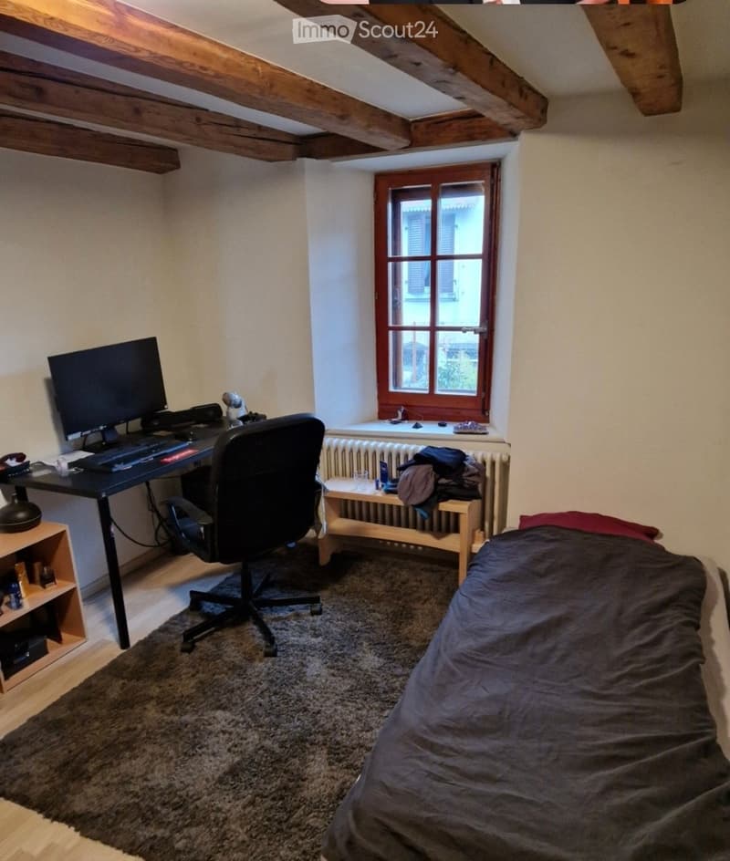A cozy bedroom with a desk, chair, and a bed, featuring exposed wooden beams on the ceiling and a window with wooden shutters.