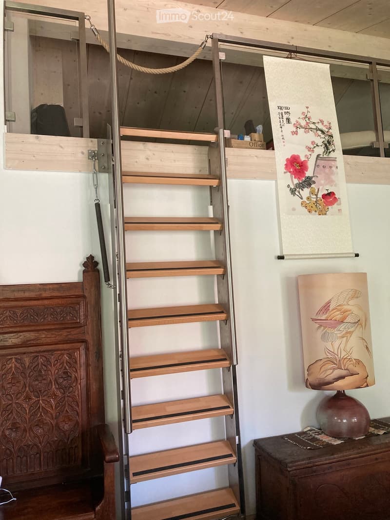 A narrow hallway with a wooden staircase, a lamp on a small table, and a painting on the wall.