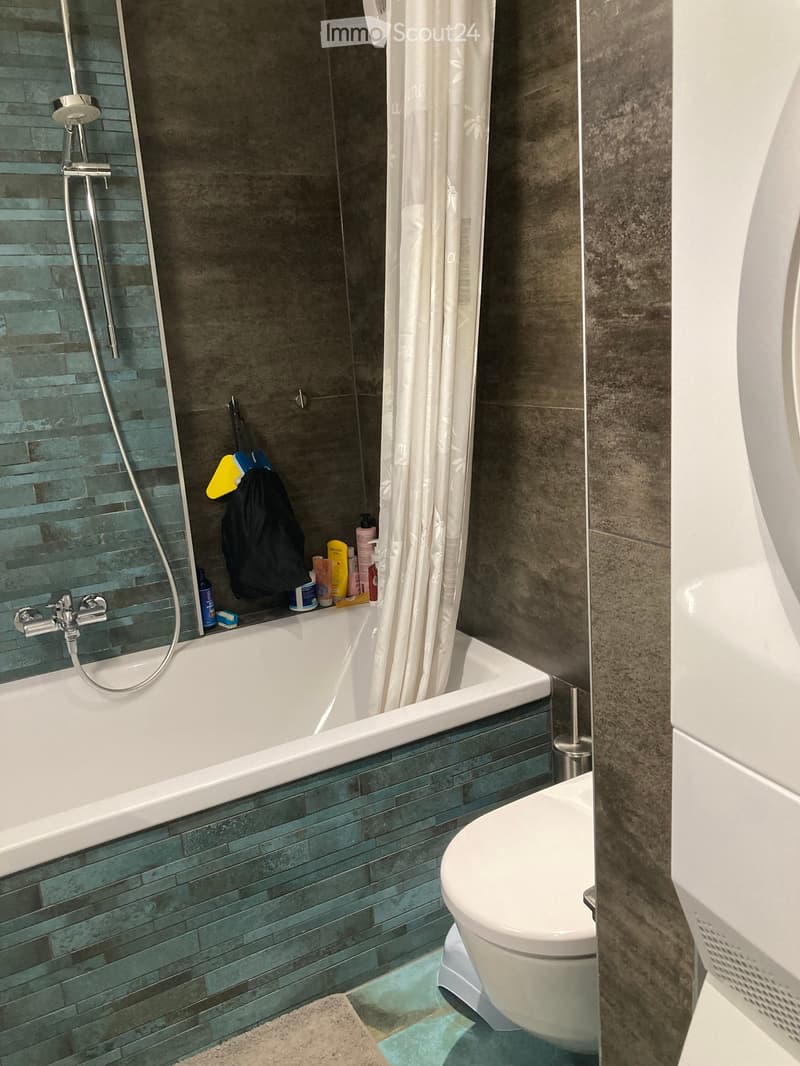 Modern bathroom with a white bathtub, toilet, and showerhead. Green tiles line the walls, and a white curtain separates the shower area. Various toiletries are placed on the edge of the bathtub.