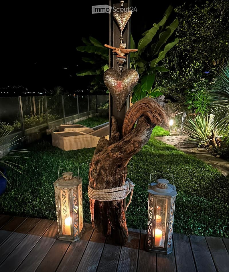 Wooden deck with lanterns, tree trunk with metal hearts, hedges, a glass fence, outdoor seating area