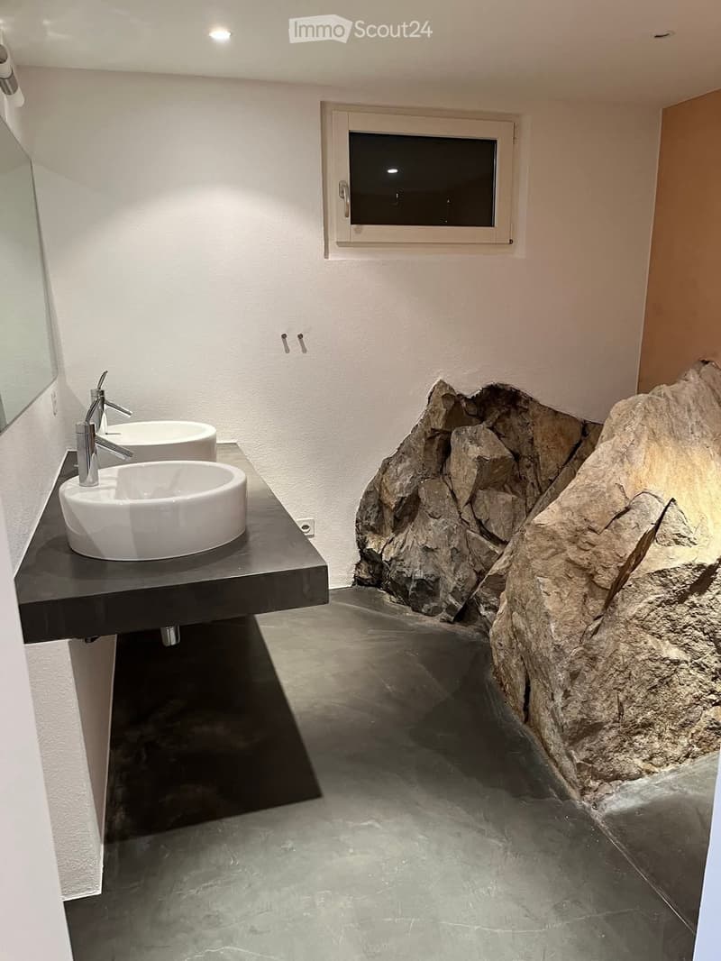 modern bathroom with concrete floors, two white basins, stone wall, mirror, no window