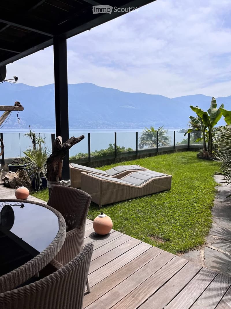Wooden patio with glass railing, deck chairs, a table, decorative items, lush green grass, and a sea view with mountains