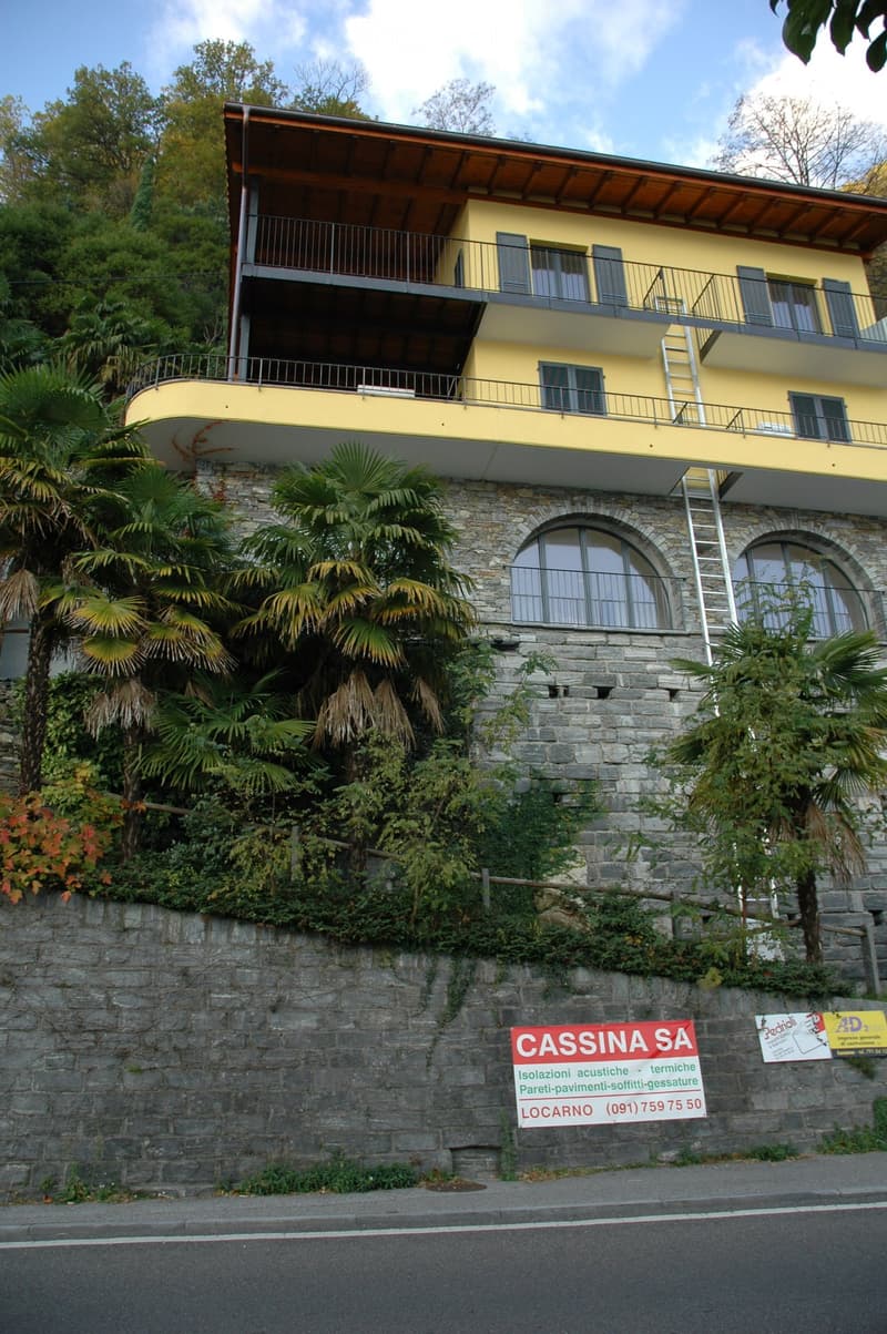 A multi-story building with a yellow exterior and balconies, set against a hillside with lush greenery.