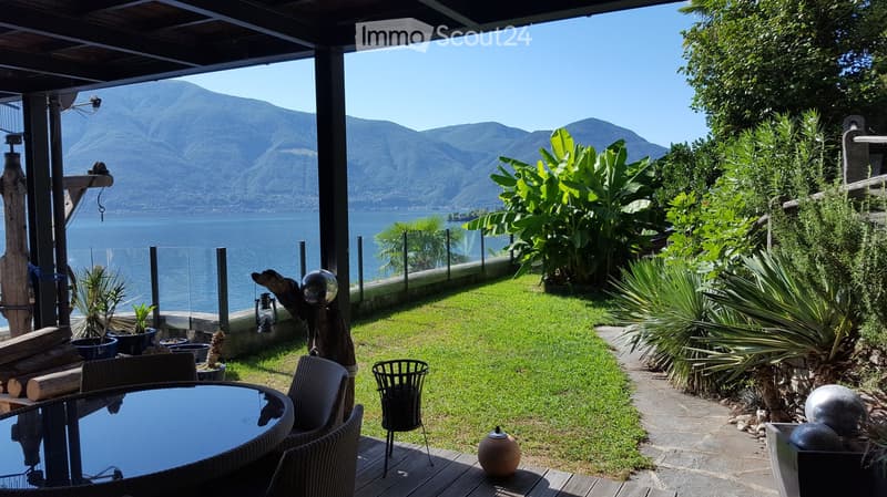 Overlooking a lake, mountains, and a fence, it has a wooden deck, a table, chairs, potted plants, and some decorative items.