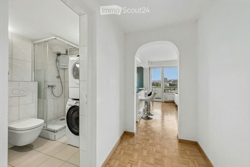 Modern hallway with wooden flooring, leading to a bathroom and a room with bar stools and a view.