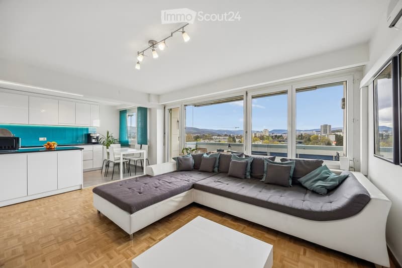 Modern, open-plan living and dining area with a large sectional sofa, coffee table, kitchen area with white cabinets and a turquoise backsplash, dining table with chairs, and large windows offering a scenic view.