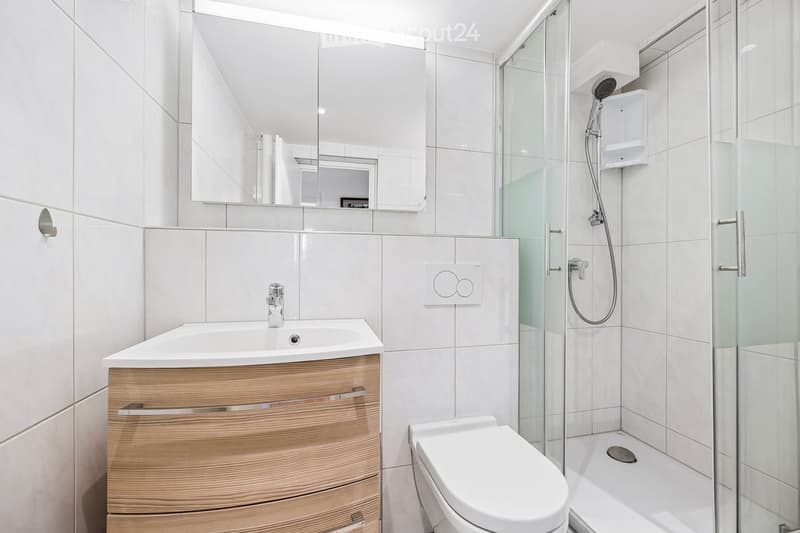 Modern bathroom with white tiles, a wooden vanity, a sink, a toilet, a shower with glass enclosure, and a large mirror above the sink.