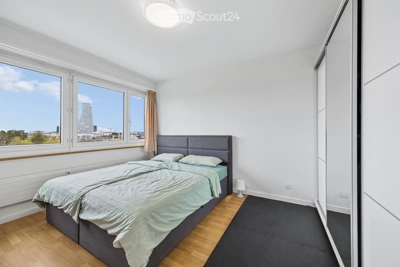 Modern bedroom with a grey headboard bed, light green bedding, wooden flooring, large windows with city view, and a small table lamp.