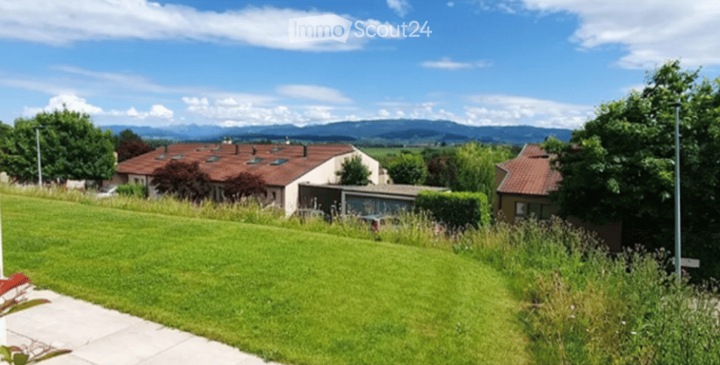 A well-maintained lawn with a pathway leading to a cluster of houses with red-tiled roofs, surrounded by trees and a scenic mountain backdrop.