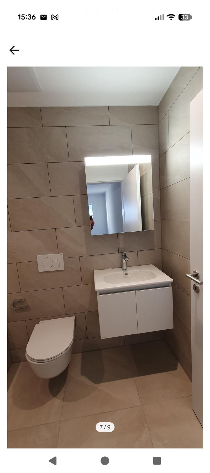 Modern bathroom with beige tiles, white toilet, sink with cabinet, and illuminated mirror above the sink.