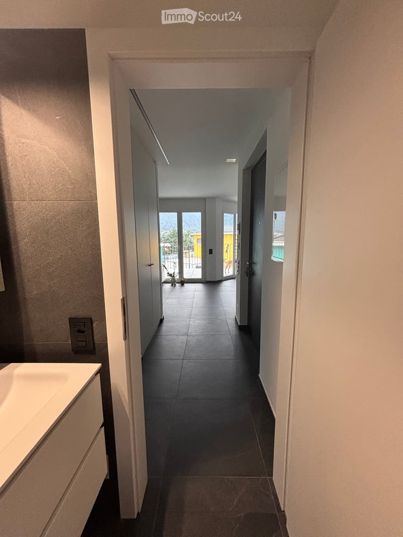Empty hallway with tiled floor, white walls, and recessed lighting. Doors lead to other rooms and an outdoor area with plants visible through a glass door.
