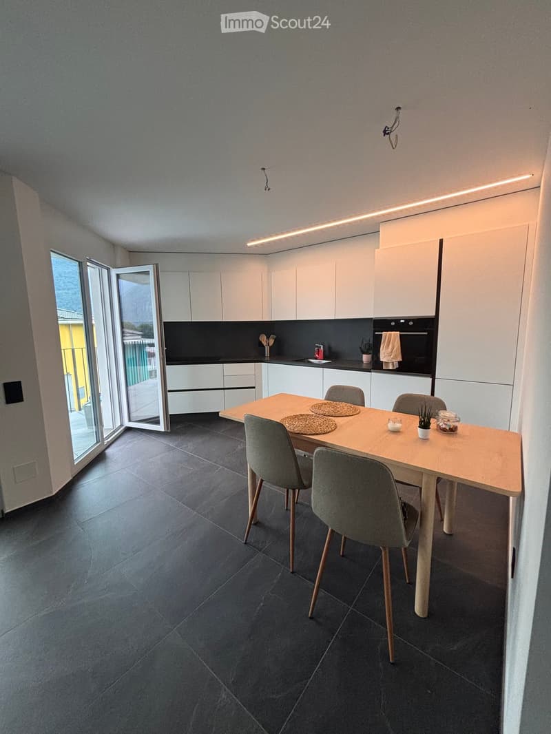 Modern kitchen with white cabinets, dark countertops, and a dining area featuring a wooden table and grey chairs. The space includes an oven, a faucet, and a small plant on the table.