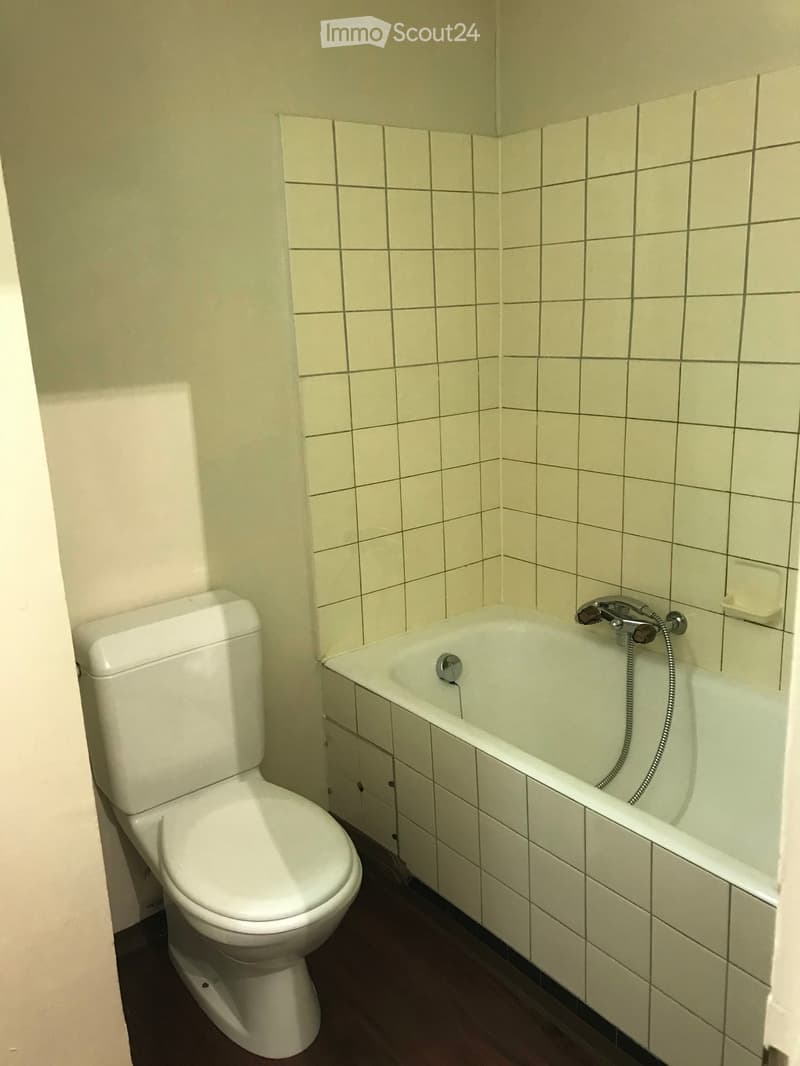 Bathroom with white tiled walls, a toilet, a bathtub with a handheld shower, and a wooden floor.