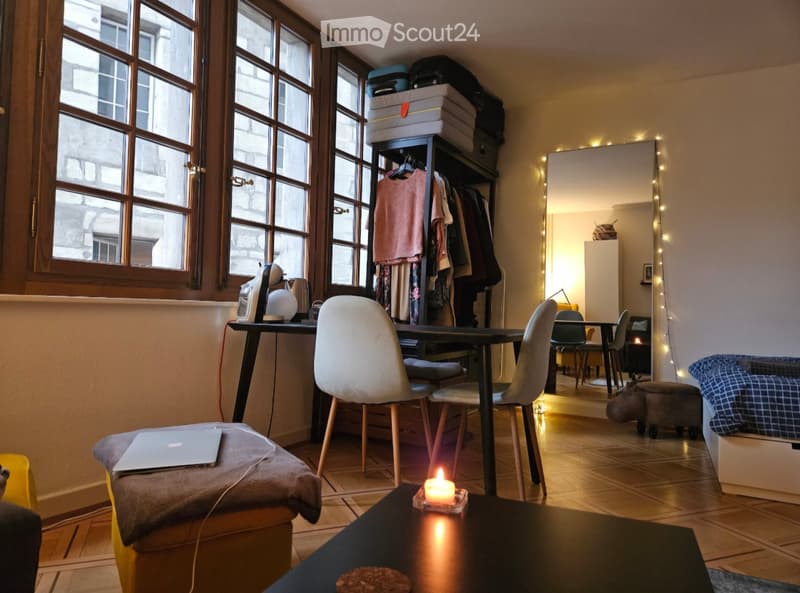 A cozy, well-lit room with large windows, a sofa, a laptop on a stool, a small table with a candle, a wardrobe, and a mirror with string lights.