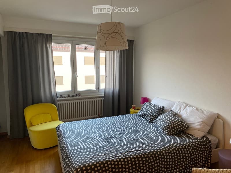 A cozy bedroom with a round bed, a yellow chair, a small table with a lamp, and a window with curtains.