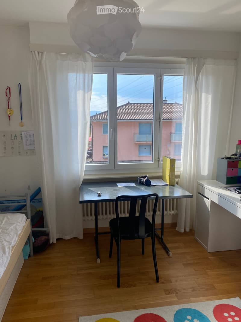 A well-lit bedroom with a wooden floor, a desk with a chair, a bed, a cabinet, and a colorful rug. The room has large windows with white curtains and a view of a pink building outside.