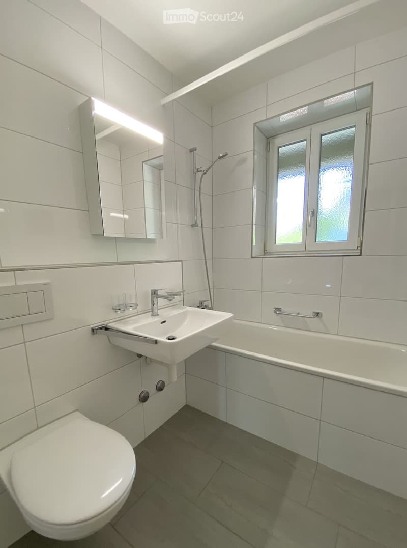 Modern bathroom with white tiles, a toilet, a sink with a faucet, a bathtub, a mirror, and a window with frosted glass.