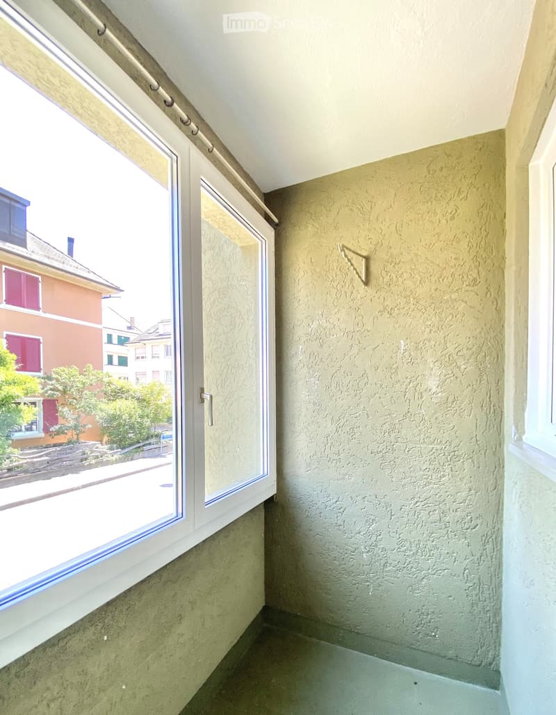 A narrow hallway with a large window on one side, showing a view of neighboring houses and greenery outside. The walls are textured and painted in a light beige color, and the floor is tiled.