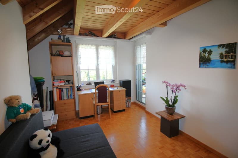 A cozy attic room with wooden floors and slanted wooden ceilings. Features include a sofa with stuffed animals, a desk with a laptop and chair, a bookshelf, a small table with a flower vase, and a painting on the wall.