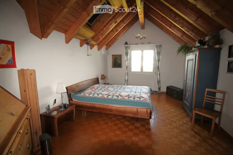 Cozy attic bedroom with wooden slanted ceilings, hardwood floors, and large window with curtains. Features a wooden bed with a colorful quilt, nightstand with lamp, wooden wardrobe, and a small chair.
