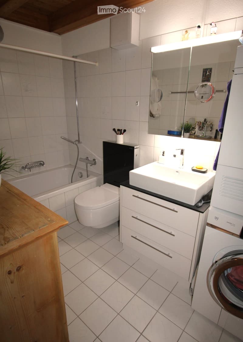 Modern bathroom with white tiles, a bathtub, toilet, sink, mirror, and a washing machine. Features include a wooden shelf and a small plant on the windowsill.