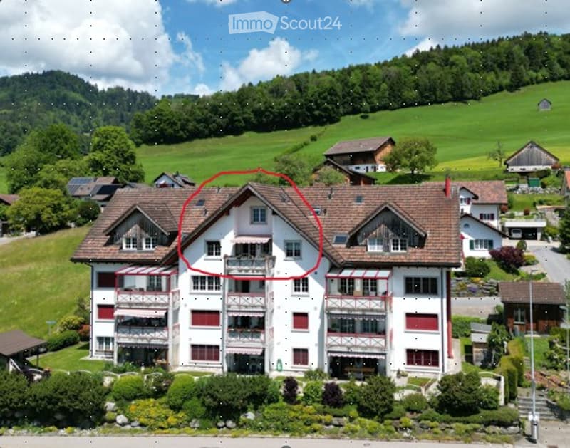 Aerial view of a multi-story residential building with white walls, red shutters, and multiple balconies, surrounded by greenery and other houses in a rural setting.