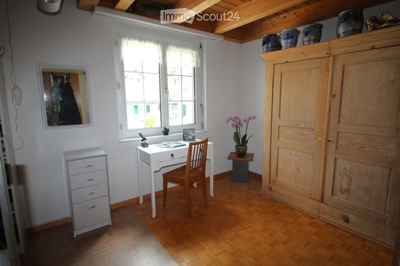 A well-lit room with wooden flooring, a large window with white curtains, a wooden chair, a white desk, a small wooden table with a flower pot, a large wooden cabinet, and a small drawer unit.